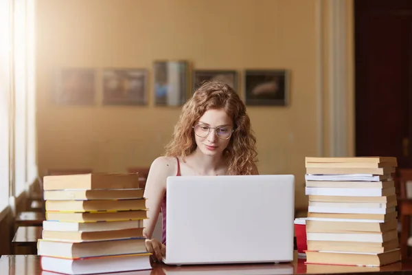 Positive tender sweet girl with curly hair working on her laptop being in reading hall with huge bunch of books on desk, focused on her studying, looking for information. Student life concept. — Stock Photo, Image