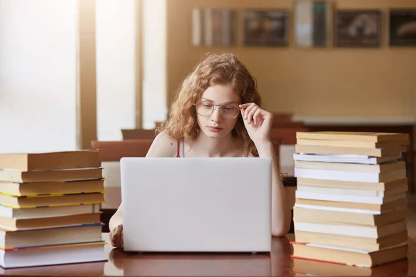 Image of serious smart college student touching her eyeglasses with one hand, looking at laptop screen attentively, searching for suitable information, being hard working student. Study concept. — Stock Photo, Image