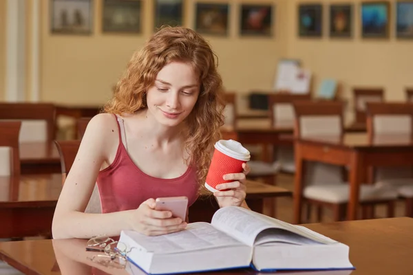 Delgado buen aspecto joven estudiante sentado en la biblioteca local, sosteniendo papercup de café y teléfono inteligente, mirando a la pantalla móvil, viendo videos, teniendo un descanso durante el estudio. Concepto de vida estudiantil . — Foto de Stock