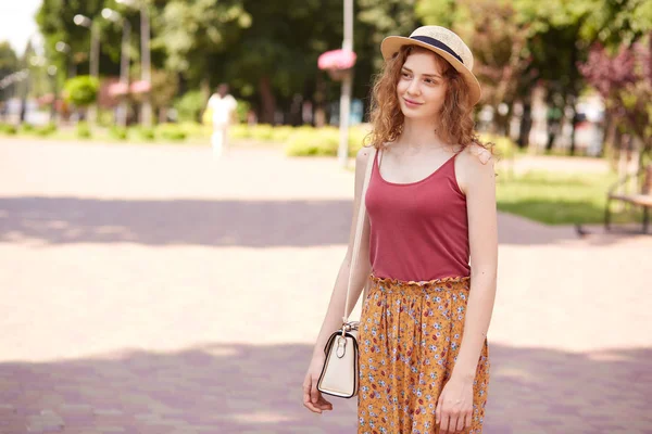 Outdoor shot of romantic adorable model with fair curly hair standing in local park, spending time around nature with pleasure, wearing hat, red top and colorful skirt, having white bag on shoulder. — Stock Photo, Image