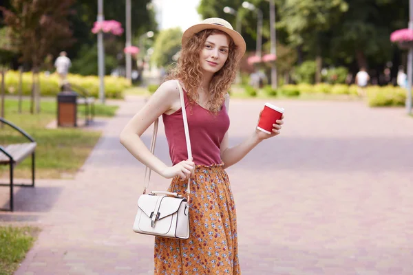 Εικόνα από ελκυστική ειλικρινή ειλικρινή κυρία με σγουρά μαλλιά που στέκονται στο μέσο του πράσινου τοπικού πάρκου, έχοντας περπατήσει, στάση για διάλειμμα για καφέ, κρατώντας κόκκινο χάρτινο κύπελλο με το ποτό. Αίσθηση εφεδρικές. — Φωτογραφία Αρχείου
