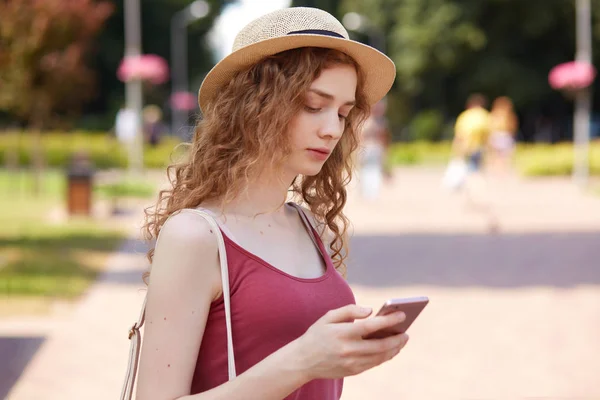 Outdoor shot of pensive cute young female holding her mobile phone in one hand, looking attentively at its screen, typing messages, checking social networking sites, spending her spare time. — Stock Photo, Image
