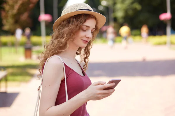 Portrait of good looking positive girl being in good mood typing messages to her friends having walk in recreation zone, holding smartphone in one hand. People and free time activities concept.