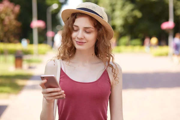 Delighted happy young lady having pleasant facial expression, smiling sincerely, reading news, holding smartphone, using it actively, wearing red shirt and fashionable summer hat. Social medias. — Stock Photo, Image