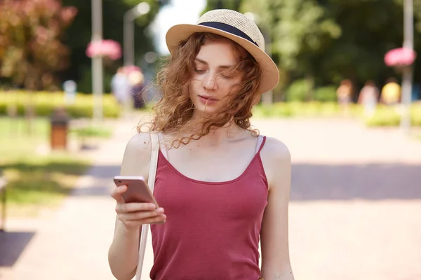 Tender sweet curly haired girl walking along green summer street, having smartphone in her hand, being focused on her mobile phone, making new post attentively, wearing casual clothes. Summer concept. — Stock Photo, Image