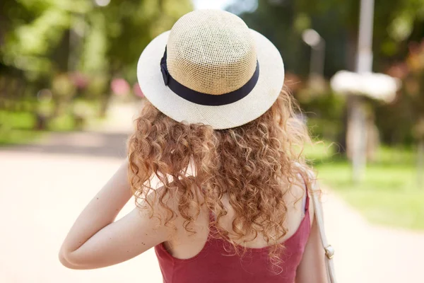 Vue arrière de la femelle élancée aux cheveux clairs et bouclés portant un chapeau de paille et une chemise rouge, touchant ses cheveux à la main, se promenant dans le parc pendant l'heure du déjeuner, aimant la nature. Concept jeunesse . — Photo