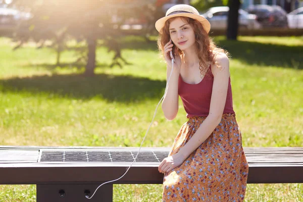 Plan extérieur de femme attrayante chargeant son téléphone sur un panneau solaire polyvalent gratuit incorporé dans le banc assis pour les citoyens, portant un chapeau, un t-shirt et une jupe au sol, regardant directement la caméra . — Photo