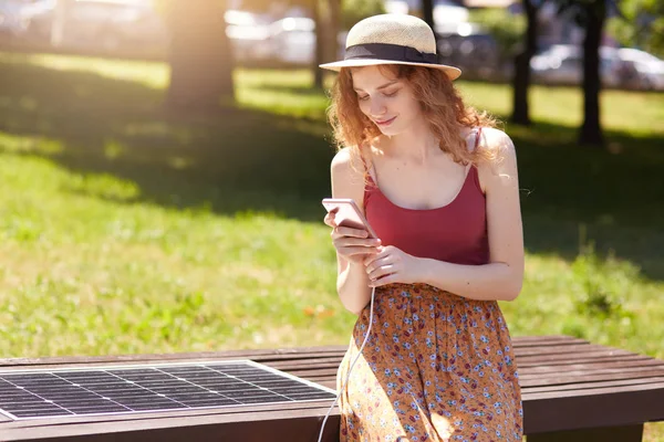 Horizontal short of young girl charges mobile phone via USB outdoors, lady sits on bench with solar panel in town park. Public charging on city street. Modern technology, ecology, alternative energy. — Stock Photo, Image