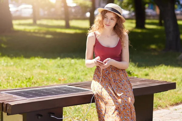 Solutions énergétiques alternatives dans la ville moderne. Jeune femme se reposant dans le parc, assis sur un banc innovant et appareil mobile de charge à partir intégré dans le port USB. Écologie, concept de technologie moderne . — Photo