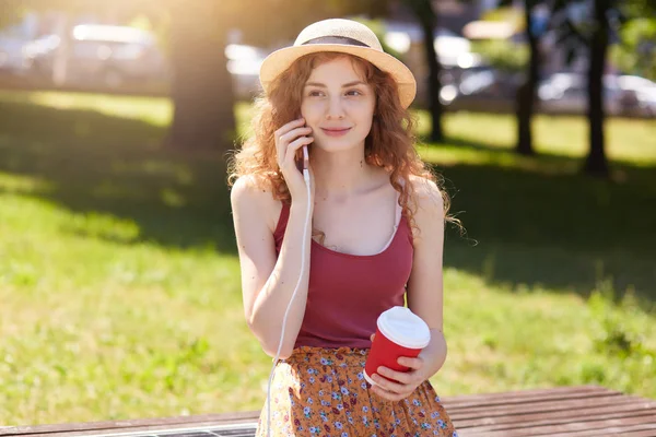 Mulher carregando telefone celular via USB em carregamento público, encantadora fêmea com cabelo ondulado foxy sentado no banco com painel solar na rua, senhora bebendo café takeaway no parque. Eletricidade alternativa . — Fotografia de Stock