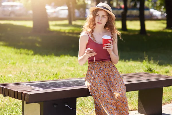 Horixontal tiro de jovem mulher carregando telefone celular enquanto sentado no banco com painel solar, fêmea tem aparência elegante, segurando xícara de plástico de café takeaway, olhando para o lado. Energia alternativa . — Fotografia de Stock