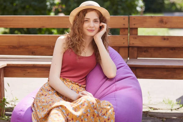 Εξωτερική βολή της νεαρής γυναίκας που κάθεται σε μεγάλο μωβ μαξιλαράκι με βάση την καρέκλα στον ανοιχτό αέρα, ελκυστική γυναίκα φορώντας casual shirt Βουργουνδίας, φούστα δαπέδου και καπέλο, έχει ξεκουραστεί με τους φίλους της στο πάρκο της πόλης. — Φωτογραφία Αρχείου