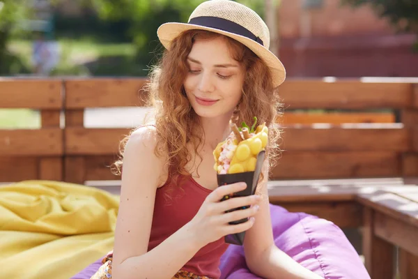 Ung kvinna sitter på farameless stol i parken, håller i händerna glass, bär vinröd casual t skjorta och hatt, ser mystiskt ner, njuter av att tillbringa sommardagen utomhus. Fritidskoncept. — Stockfoto