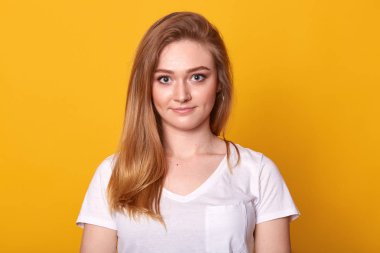 Manyetik çekici kadın sarı arka plan üzerinde izole poz Stüdyo çekim, doğrudan kameraya bakarak, mavi büyük gözleri ve uzun adil saç sahip, rahat beyaz t shirt giyiyor. İnsan kavramı.