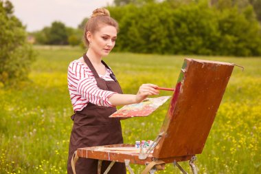 Resim çizen genç kadının görüntüsü, doğada çizim için eskiz defteri ni kullanan, ressam kız fırça ve renk paleti ile duruyor, konsantre görünüyor, çayır manzara boyama. Sanat kavramı.