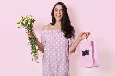 Gülümseyen genç Avrupalı kadının Stüdyo çekim koyu uzun dalgalı saçları vardır, polka nokta gül elbise giyen, hediye çantası ve çiçek buketi tutar, açık pembe stüdyo arka plan üzerinde poz, doğum günü var.