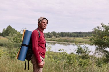 Sırt çantası ve mavi halı ile yaşlı adam Yan görünümü, onun aktif tatil sırasında harika kırsal yürürken nehir hayran görkemli görünümü zevk. Seyahat ve sırt çantası konsepti.