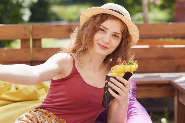 フレームレスチェアの上に座っている若い女性の肖像画、アイスクリームを食べて、夏の公園で自分撮り写真を撮って、大学の試験に合格した後にリラックスし、スタイリッシュな服と帽子を身に着けている. — ストック写真