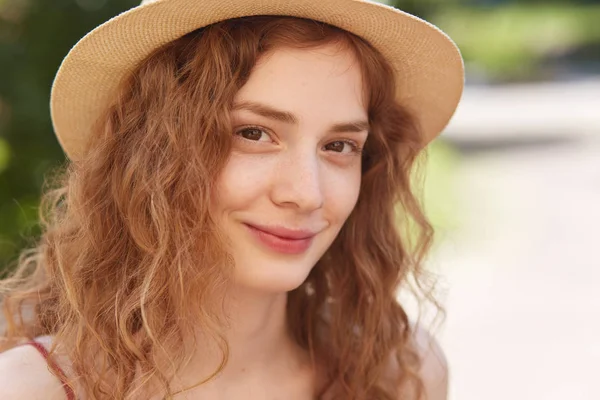 Primer plano retrato al aire libre de la joven chica atractiva con sombrero de paja, tiene el pelo ondulado foxy, mirando directamente a la cámara, tiene una sonrisa encantadora, posando en el parque de la ciudad, se ve tranquilo y feliz. Concepto de personas . —  Fotos de Stock