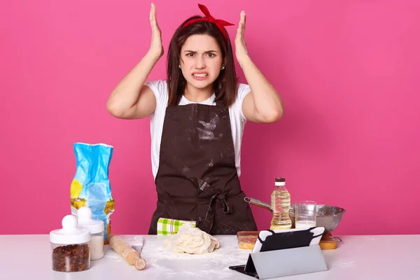 Horizontal shot of angry housewife, lady wearing brown apron dirty with flour, white casual t shirt, red headband, keeps hands up, looks tired, model posing isolated over pink studio background.