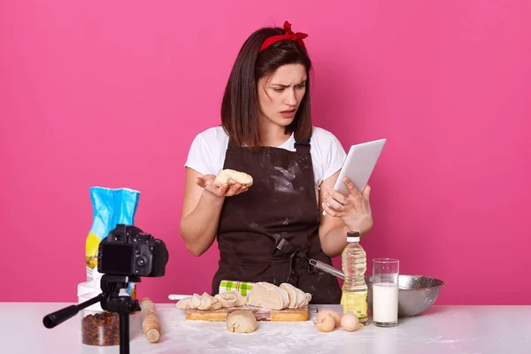 Portrait of cooking amateur holding dough in one hand and tablet, watching culinary lessons, confused with recipe, having surprised facial expression, wearing headband, dirty apron and white t shirt.