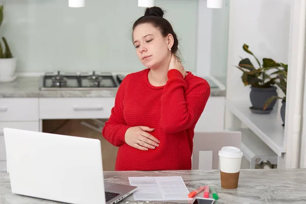 Indoor shot of tired Caucasian woman keeps hand on neck, working in kitchen at laptop computer, wearing casual red sweater, anticipates for baby, poses against kitchen interior, earning money online. — Stock Photo, Image