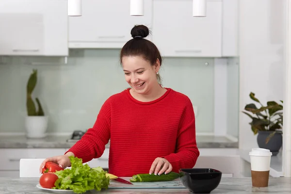 La foto horizontal de la feliz mujer embarazada sonriente corta verduras para hacer ensalada, posa contra el interior de la cocina, usa suéter casual rojo, linda mujer espera al niño. Concepto de maternidad e hijos — Foto de Stock