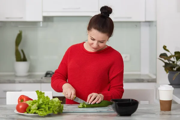 Inyección interior de mujer embarazada cocinando en casa, haciendo ensalada verde fresca para la cena, comiendo muchas verduras diferentes durante el embarazo, concepto de embarazo saludable, señora con suéter casual rojo . — Foto de Stock