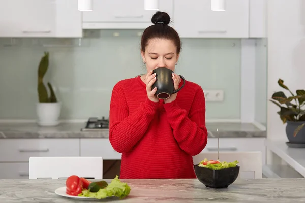 Close up portrait of good looking woman keeps cup of tea or coffee with both hands, drinks hot beverage, pregnant female poses against kitchen interior, enjoys calm atmosphere. Motherhood concept. — Stock Photo, Image