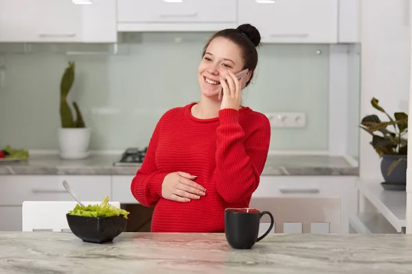 Фото счастливой смеющейся брюнетки, сидящей на кухне и разговаривающей по смартфону с подругой или мужем, женщина ждет ребенка. Беременная женщина проводит время дома. Технологическая концепция . — стоковое фото