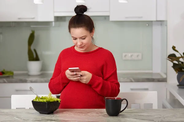 Imagen de una mujer embarazada saludable increíble en la cocina en casa usando el teléfono móvil para comprobar la red social o correo electrónico, comer ensalada y beber té o café. Pragnancy y concepto de tecnología moderna . — Foto de Stock