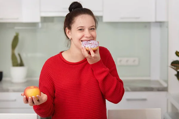 Mujer embarazada comiendo pastel dulce y sosteniendo la manzana en otra mano, se pone en pie vistiendo suéter casual rojo en el frente interior de la cocina blanca, mujer expresa felicidad y alegría. Concepto de pragnancia . — Foto de Stock