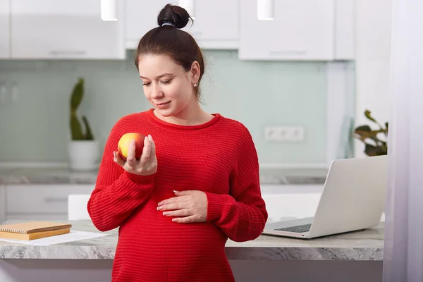 Pregnant beautiful girl holding apple in hand. Attractive future mother feels relaxed and happy, charming woman has good health, takes good care of herself and her baby by eating healthy foods. — Stock Photo, Image