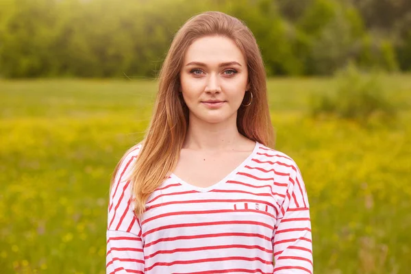 Outdoor portrait of happy smiling girl with long straight hair in white shirt with red stripes posing in summer meadow, has calm and pleasant facial expressions, being photographed by friend. — Stock Photo, Image