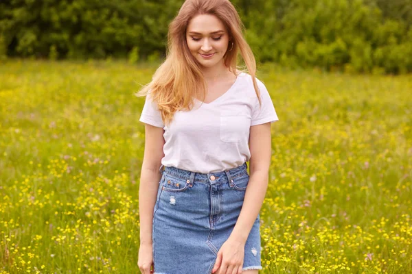 Imagem de menina branca bonita jovem em t-shirt branca e saia jeans azul, posando no prado e olhando para baixo, tem cabelo loiro longo estranho, expressa alegria e felicidade. Conceito de pessoas . — Fotografia de Stock