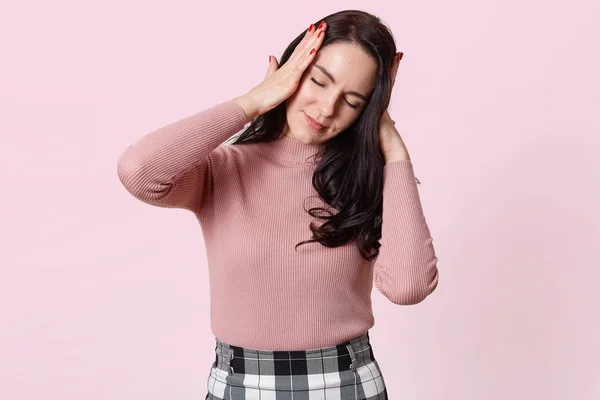 Studio shot di attraente giovane donna con terribile mal di testa, tiene entrambe le mani sulla testa, ha bisogno di prendere la medicina, bruna femmina in camicia rosa e gonna a scacchi, isolato su sfondo rosa . — Foto Stock