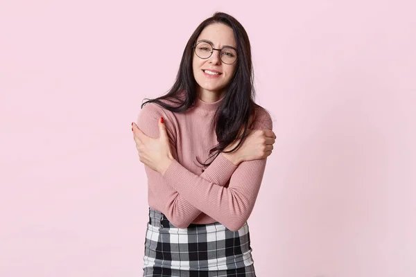 Retrato de primer plano de atractiva mujer guapa, abrazándose a sí misma a causa del frío, vistiendo suéter rosa, falda y gafas con estilo, temblorosa, dentadura fija, aislada sobre fondo rosa . —  Fotos de Stock
