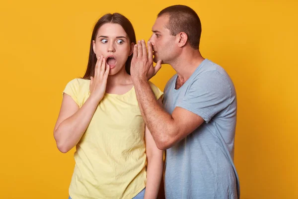 Man covering his mouth and whispering secret to girlfriend ear, brunette emotional young woman looking shocked, opening eyes and mouth widely with surprise, isolated over yellow background in studio.