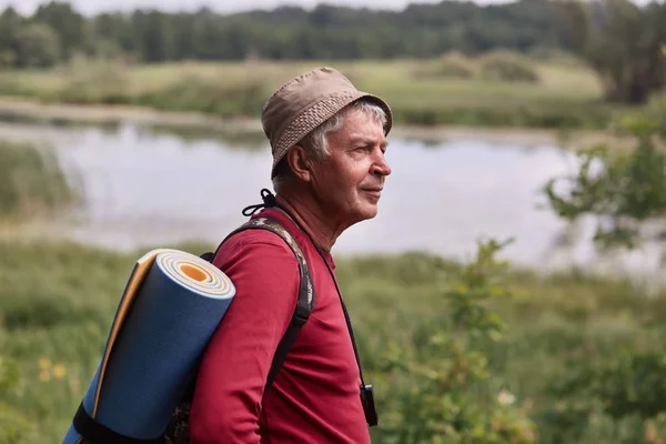 Profilo colpo di uomo anziano con zaino e tappeto blu, indossando maglione casual rosso e cappello, godendo bellanatura vicino al fiume e guardando lontano. Zaino in spalla e concetto di ricreazione attiva . — Foto Stock