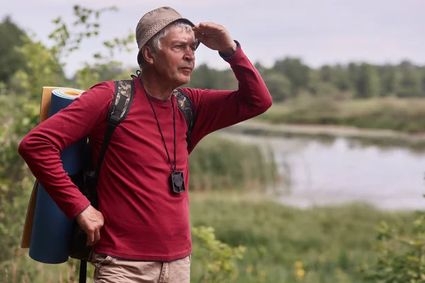 Foto di uomo caucasico hipster uomo con zaino e tappeto in fondo alla natura. Tempo di relax in vacanza, maschio anziano guardando lontano, vestita casual camicia rossa e cappello. Viaggiare e riposo attivo . — Foto Stock