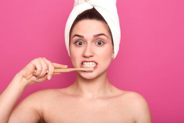 Banyoda diş fırçası ve diş macunu ile dişlerini fırçalayan, çıplak omuzlar ve sarılmış havlu ile sabah prosedürleri yapıyor çekici kadın, pembe üzerinde izole, yüz ifadesi şaşırttı.