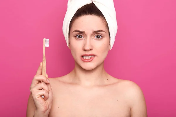 La foto interior de una mujer molesta mordiéndose el labio y sosteniendo el cepillo de dientes aislado sobre el fondo rosa del estudio, posando con hombros desnudos y una toalla blanca envuelta en la cabeza, se ve descontenta. Concepto de higiene . — Foto de Stock