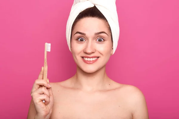 Portret van glimlachend mooie vrouw met verbasterd gezichtsuitdrukking, houdt tandenborstel geïsoleerd over roze achtergrond en kijken direct naar camera, poseren met Nacked schouders en witte handdoek op hoofd. — Stockfoto