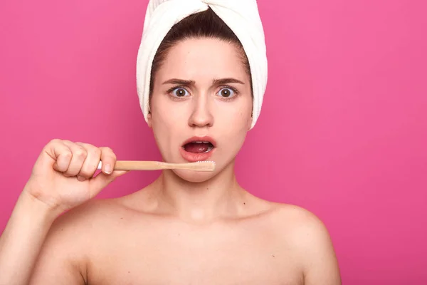 Primer plano retrato de la señora sorprendida con cepillo de dientes en el baño, cepilla los dientes por la mañana, posa con hombros desnudos y toalla blanca en la cabeza, aislado sobre fondo rosa, se ve asombrado o sorprendido — Foto de Stock