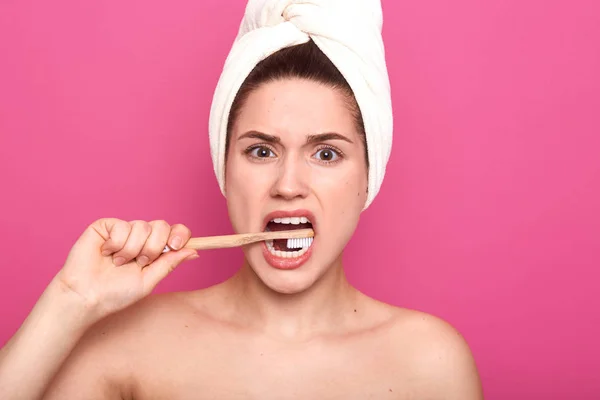 Estudio interior plano de mujer joven enérgica disgustado mirando directamente a la cámara, cepillarse los dientes intensamente con cepillo de dientes, estar desnudo con toalla en la cabeza, de pie aislado sobre fondo rosa . — Foto de Stock