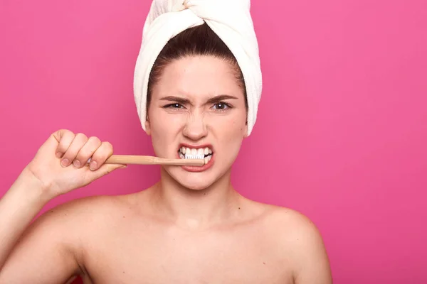 Retrato de cerca de una señora divertida posando desnuda, sosteniendo su equipo dental en una mano, cepillándose los dientes cuidadosamente, cubriéndose el cabello con toalla blanca, de pie aislada sobre fondo rosa . — Foto de Stock