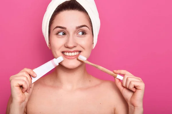 Mujer soñando en el baño, atractiva hembra con cepillo de dientes y pasta de dientes en las manos posando aislada sobre fondo rosado, mirando a un lado, toalla blanca envuelta, procedimientos matutinos en el baño . — Foto de Stock