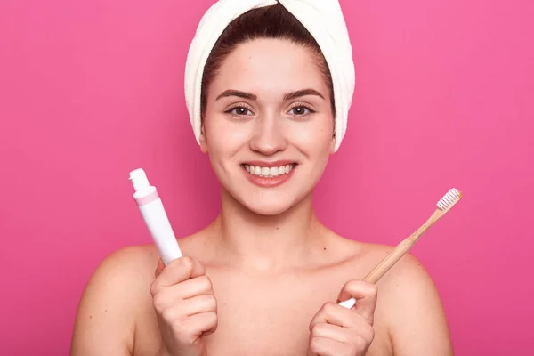 La foto horizontal de la encantadora mujer caucásica sonriente aislada en la pared del estudio rosado, lista para cepillarse los dientes, sosteniendo la pasta de dientes y el cepillo de dientes en las manos, tiene una piel suave y perfecta. Concepto de higiene . — Foto de Stock