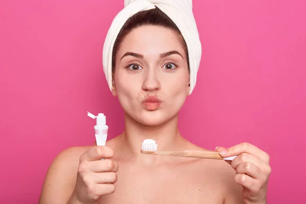 Indoor shot van tevreden vrouw met afgeronde lippen, model maakt Kiss gebaar, Pouts lippen, draagt witte handdoek op hoofd, heeft douche, poseren met tandenborstel en tandpasta. Hygiëne en Beauty concept. — Stockfoto