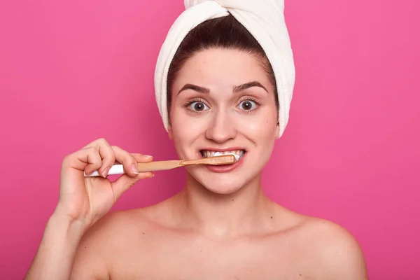 Close up shot of girl with funny facial expression, posing with toothbrush, young female cleans her teeth, standing in bathroom with towel on head, isolated over pink background. Oral hygiene concept.